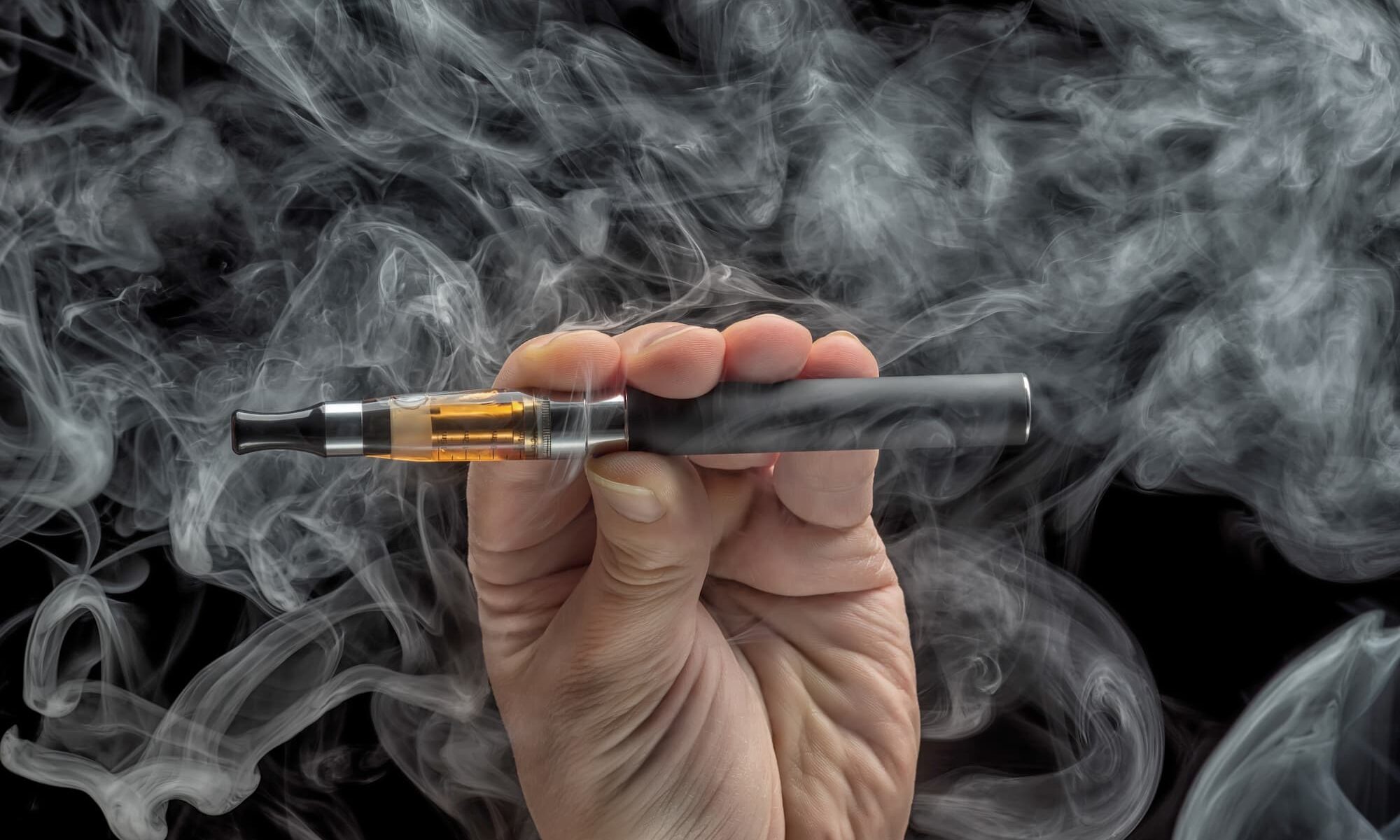 A close up image of a hand holding a vape pen with clouds of smoke in the background